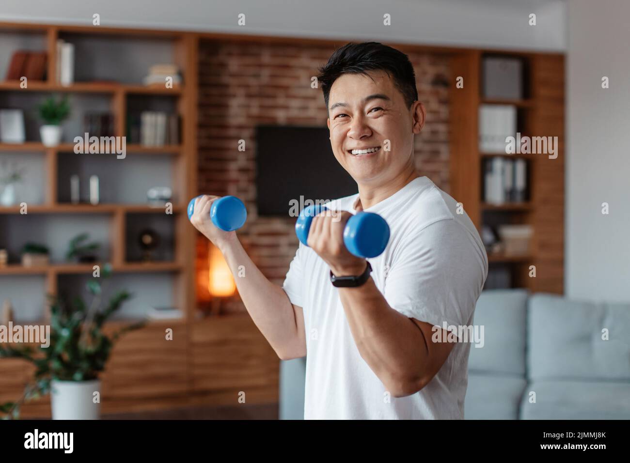 L'uomo maturo coreano allegro che fa esercizi di forza per le braccia, sollevando manubri in salotto, sorridendo alla macchina fotografica Foto Stock