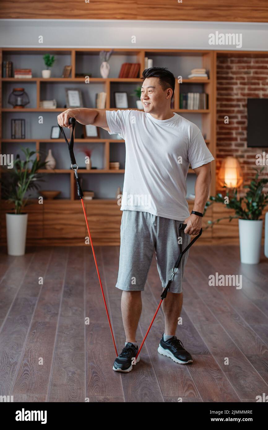 Uomo maturo asiatico sportivo esercitandosi con la fascia di resistenza nel paese, avendo workout domestico nell'interno del soggiorno Foto Stock