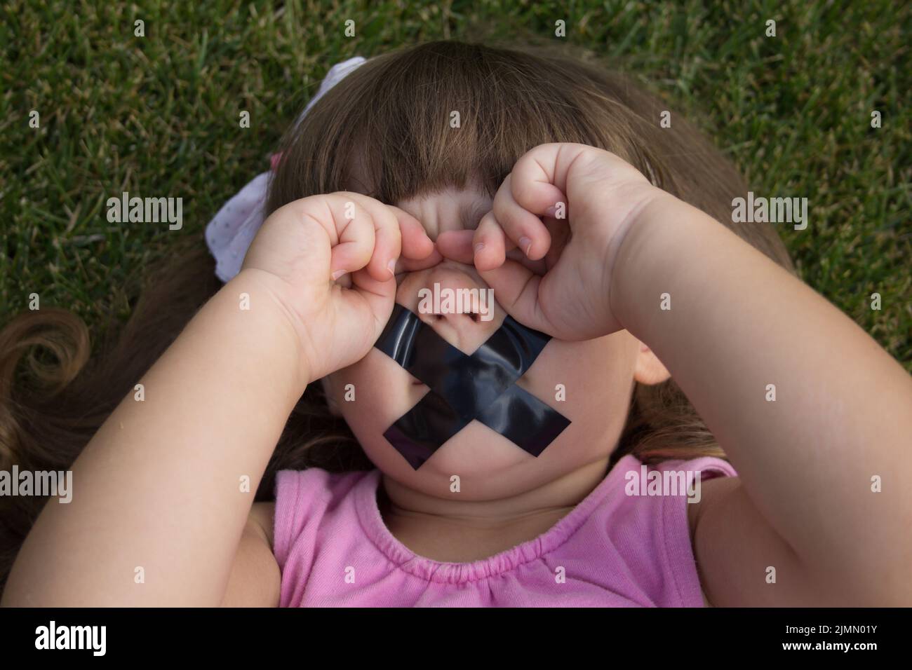 Immagine di una bambina adorabile che gli sfrega gli occhi e ha nastro elettrico per chiudere la bocca. Foto Stock