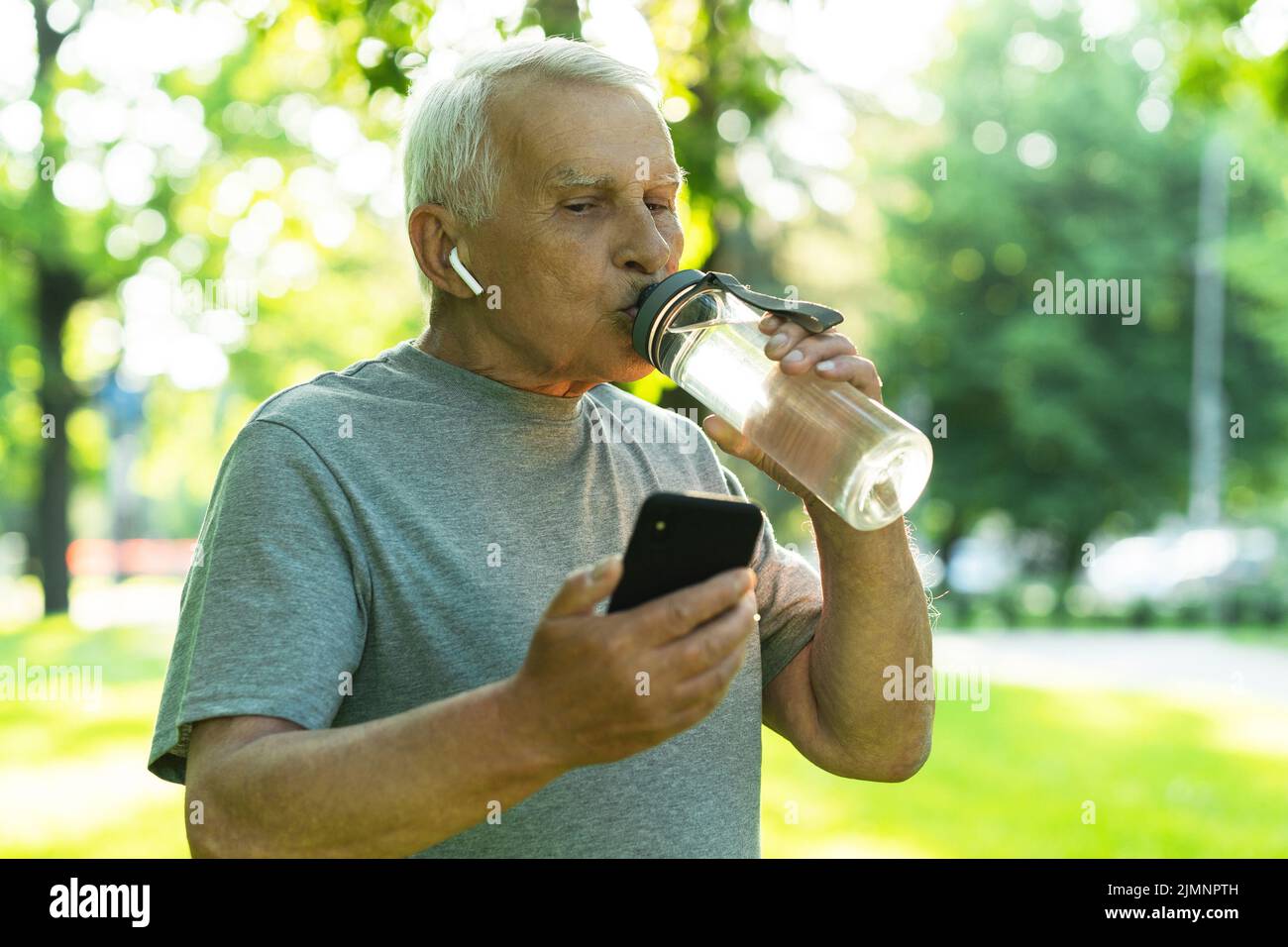 Felice uomo anziano attivo con una bottiglia d'acqua e smartphone nel verde del parco cittadino Foto Stock