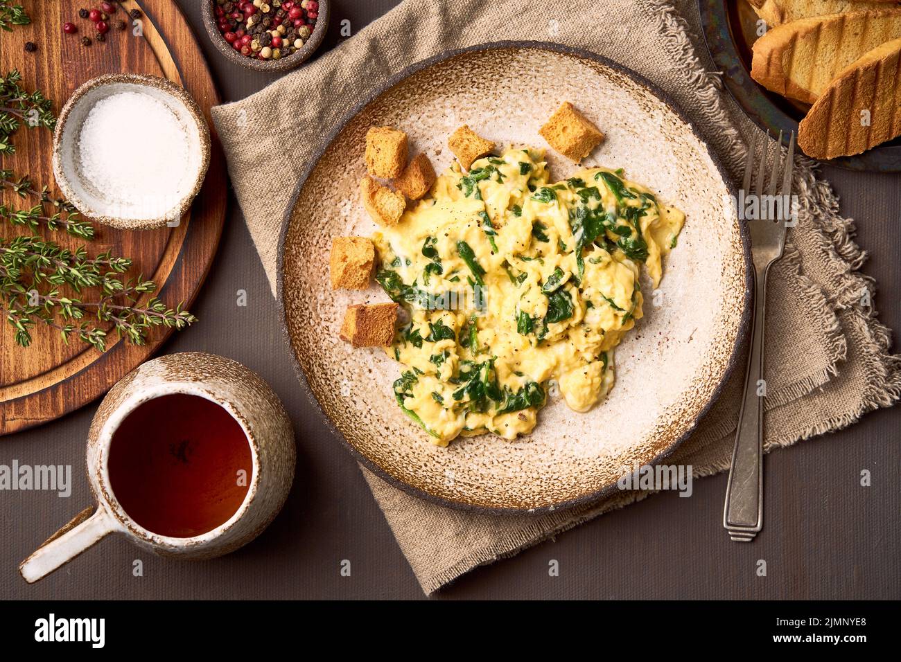 Uova strapazzate e spinaci fritti in padella, tazza di tè su fondo marrone scuro Foto Stock