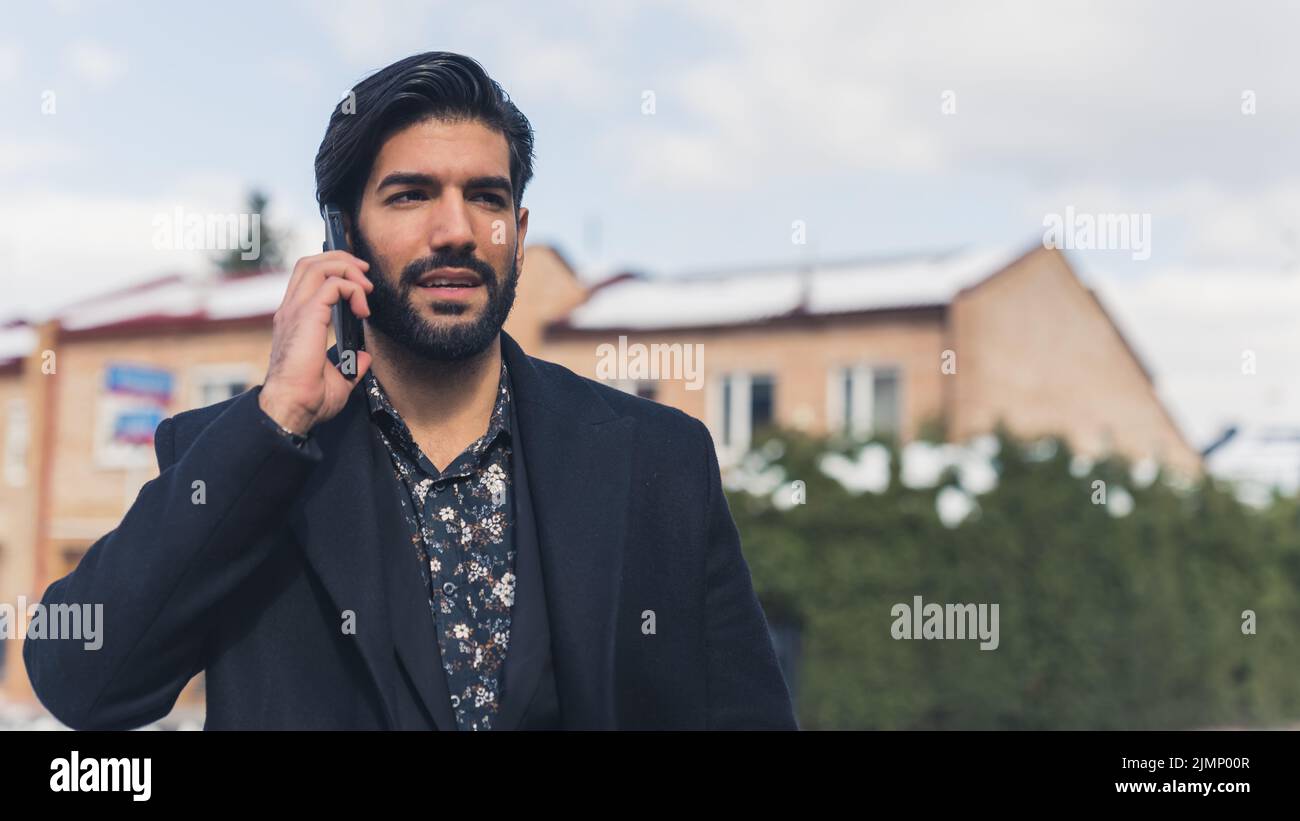 Un uomo ispanico con capelli lisci e barba che indossa una camicia e un cappotto che cammina per la città parlando al telefono. Chiamata di lavoro. Foto di alta qualità Foto Stock