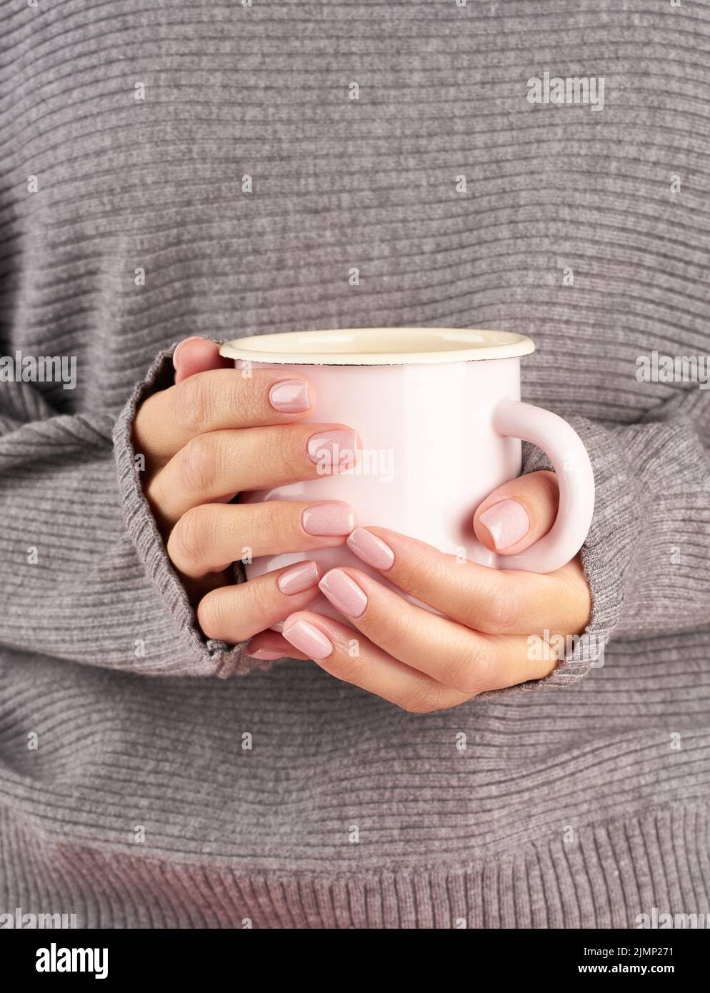 Caffè caldo mattutino al lavoro in una fredda mattinata autunnale, mani in mano con una tazza con una bevanda, maglione grigio, manicure rosa, primo piano, ve Foto Stock