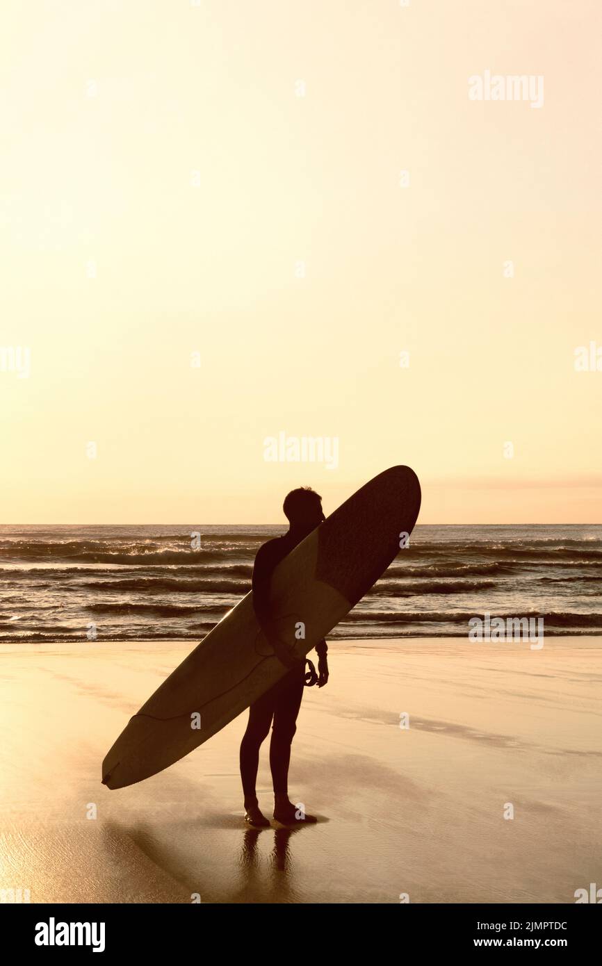 Silhouette di persona irriconoscibile con surf sulla spiaggia al tramonto. Retroilluminazione. Attività acquatiche all'aperto. Spazio per il testo. Foto Stock