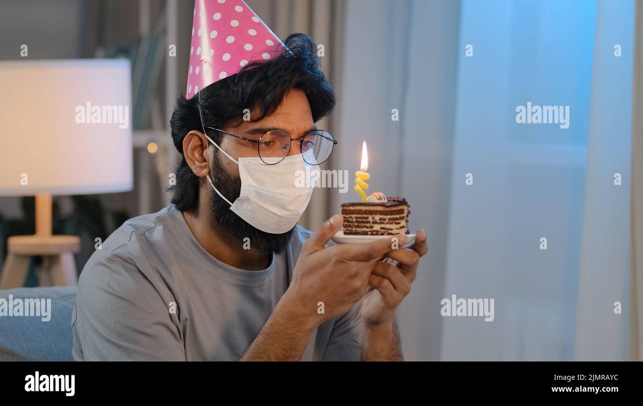 Ritratto arabo malato malato indiano bearded triste uomo in maschera medica seduta da solo a casa indossa festive compleanno rosa cappello e bicchieri tiene cupcake Foto Stock