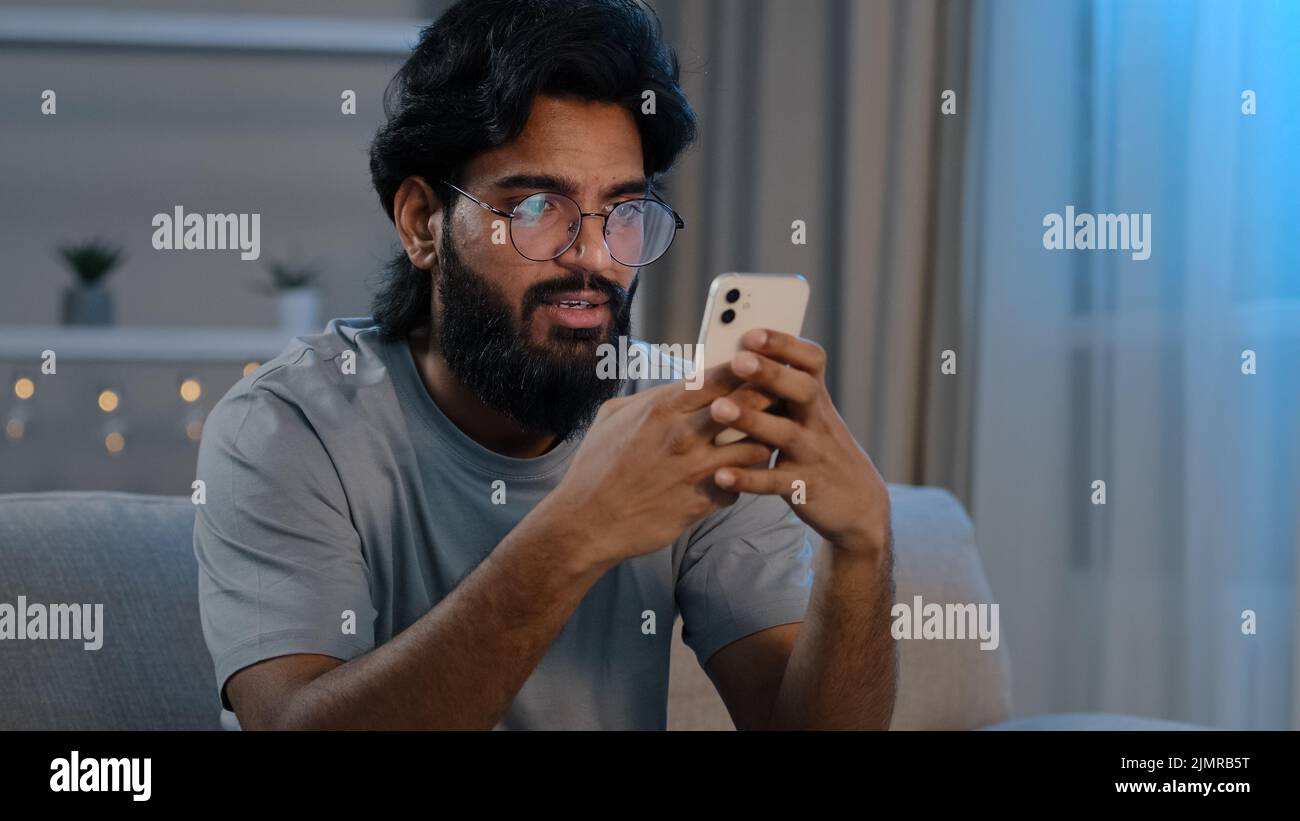 Grave uomo arabo indiano occupato bearded maschio in occhiali digitando navigazione in smartphone SMS chat utilizzando la messaggistica app mobile a casa su Foto Stock