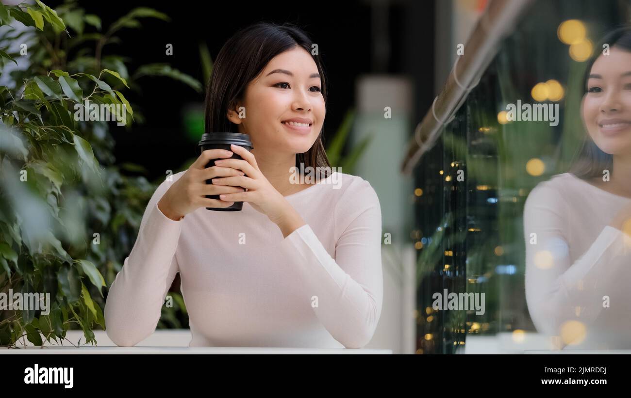 Ragazza asiatica donna sorridente godendo di bere fragrante caffè delizioso caldo tè mattina cappuccino in caffè seduto al tavolo sorridente in attesa di riunione Foto Stock