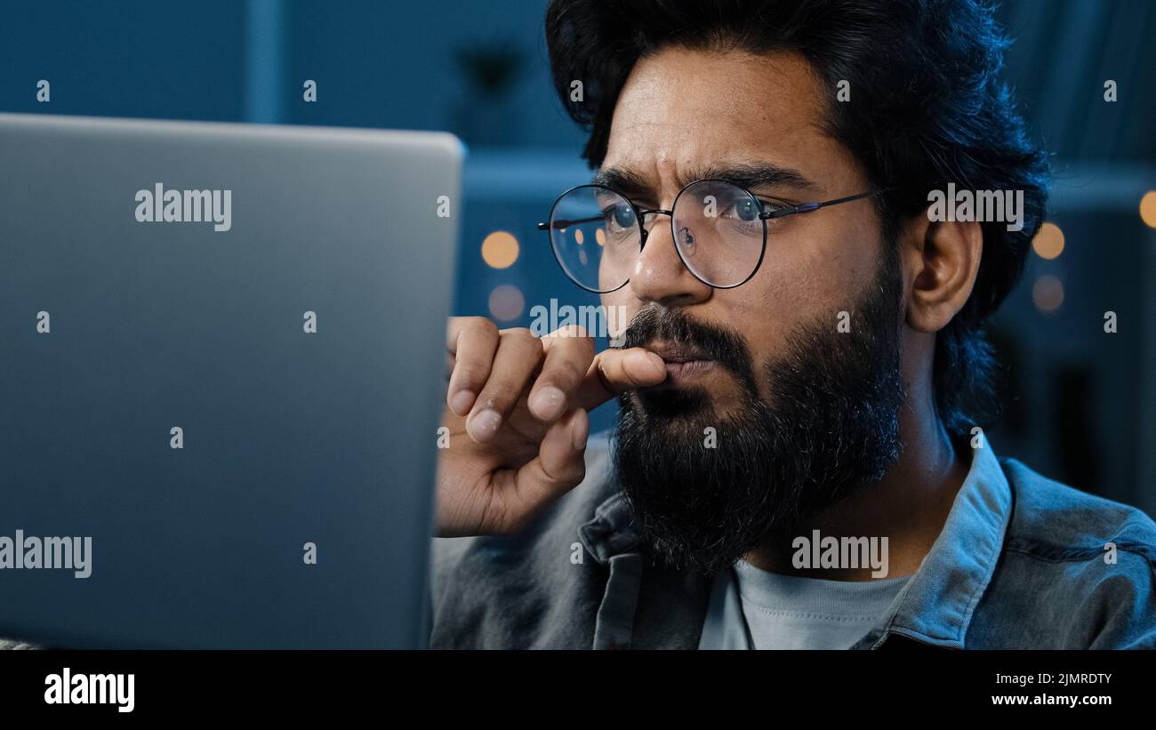 Messo a fuoco indiano barbuto uomo freelancer in occhiali che lavorano sul computer a casa tarda notte guardando la decisione di ricerca schermo sente dubbio Foto Stock