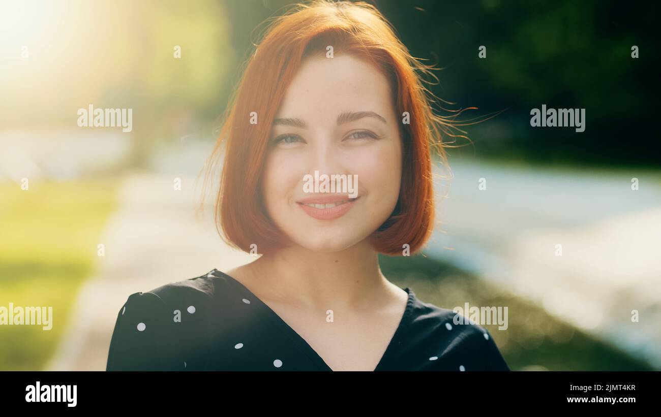 Ritratto di felice attraente femmina viso rosso elegante ragazza con acconciatura breve sorridente in posa in città raggi solari invia aria bacio a mano gesto Foto Stock