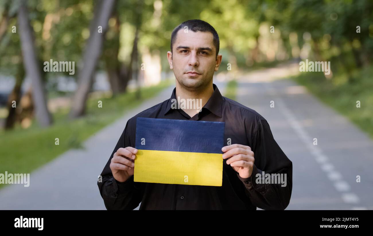 Uomo serio caucasico adulto patriota attivista manifestante indossare camicia nera stand fuori nel parco tenere disegno di cartone con bandiera nazionale blu giallo di Foto Stock