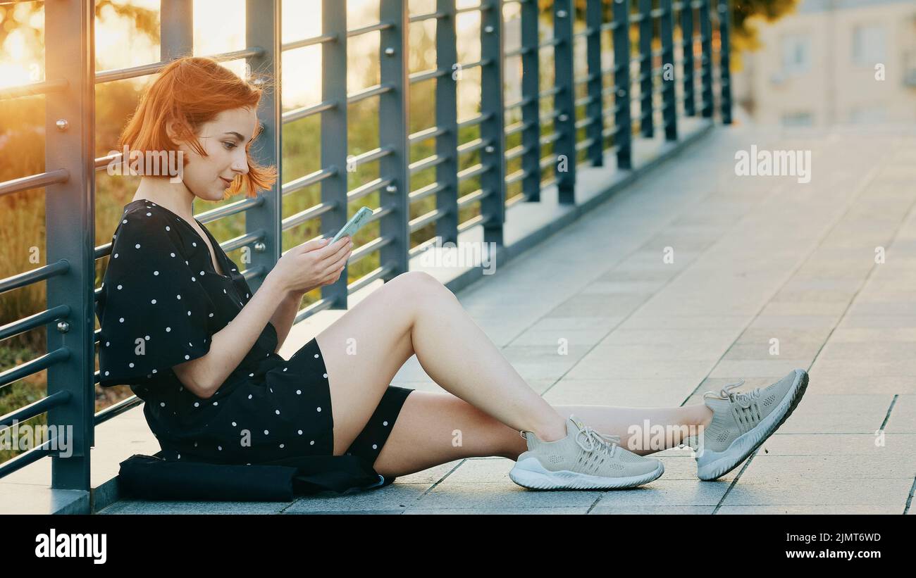 Attraente giovane ragazza adolescente bella donna rossa con capelli corti sedersi sul ponte della città resto all'aperto guarda schermo mobile guardare divertente video internet Foto Stock