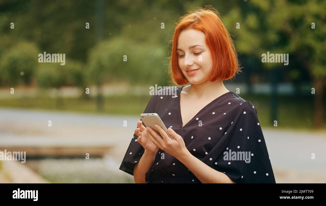 Bella rossa focalizzata caucasica ragazza adolescente donna utente con capelli corti stand in città parco tenere dispositivo moderno smartphone gadget guardare mobile Foto Stock