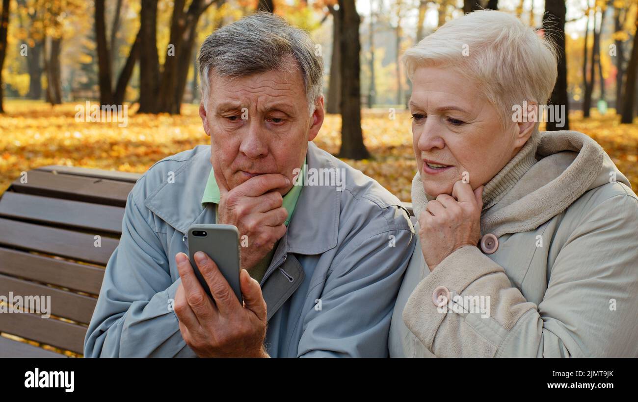 Sconvolto anziano coppia sposata sedersi in panchina in autunno parco leggere cattive notizie sullo smartphone preoccupato persone anziane guardare schermo telefono triste famiglia sensazione scioccante Foto Stock