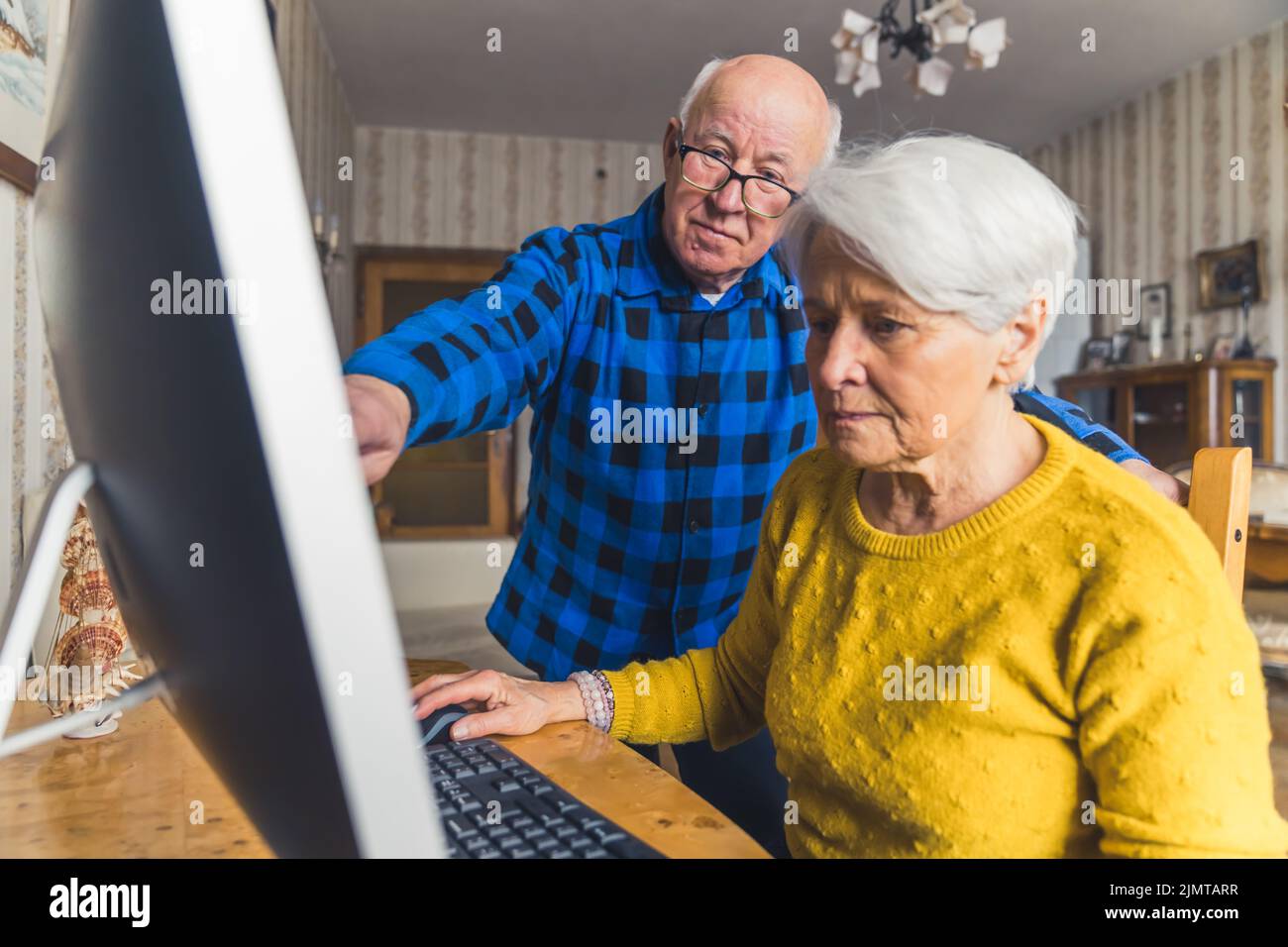 Uomo anziano e moderno esperto in piedi, che punta al monitor e insegna a sua moglie vecchia come usare il computer . Foto di alta qualità Foto Stock