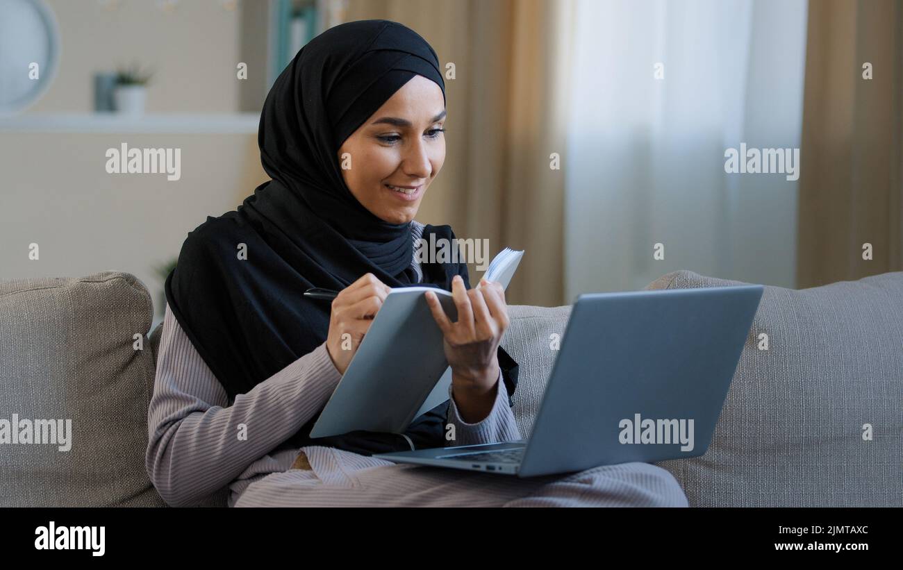 Allegro giovane musulmana signora araba giovane donna seduta sul divano accogliente scrittrice freelance femmina prendere appunti in diario notando informazioni scrivere libro compiti quotidiani Foto Stock