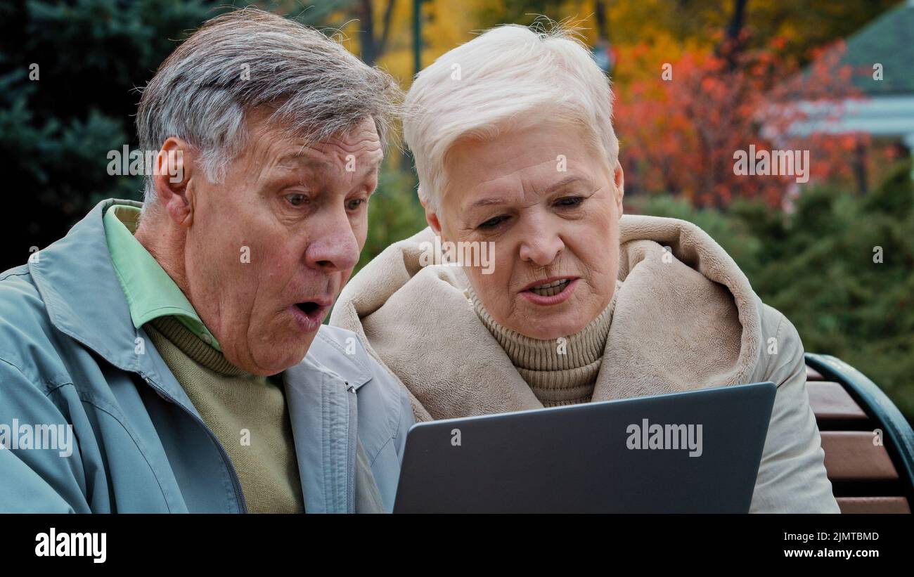 Scioccato sconvolto anziano sposato coppia partner guardando il notebook schermo sedersi in autunno parco deluso vecchia donna leggere cattive notizie preoccupata famiglia matura Foto Stock