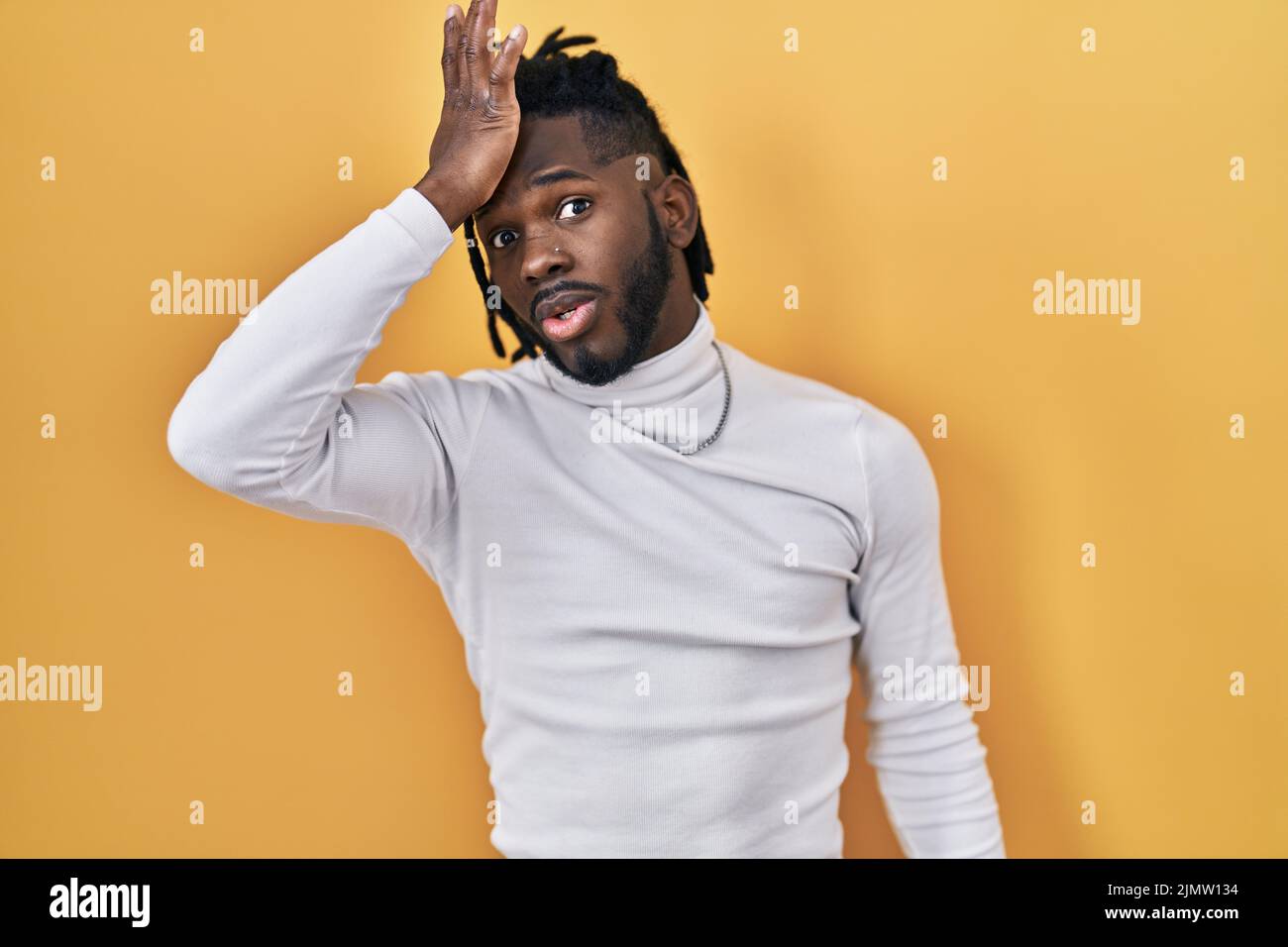 L'uomo africano con i doadlocks che indossano il maglione del dolcevita sopra lo sfondo giallo sorpreso con la mano sulla testa per errore, ricordisi di errore. dimenticato, Mem difettoso Foto Stock
