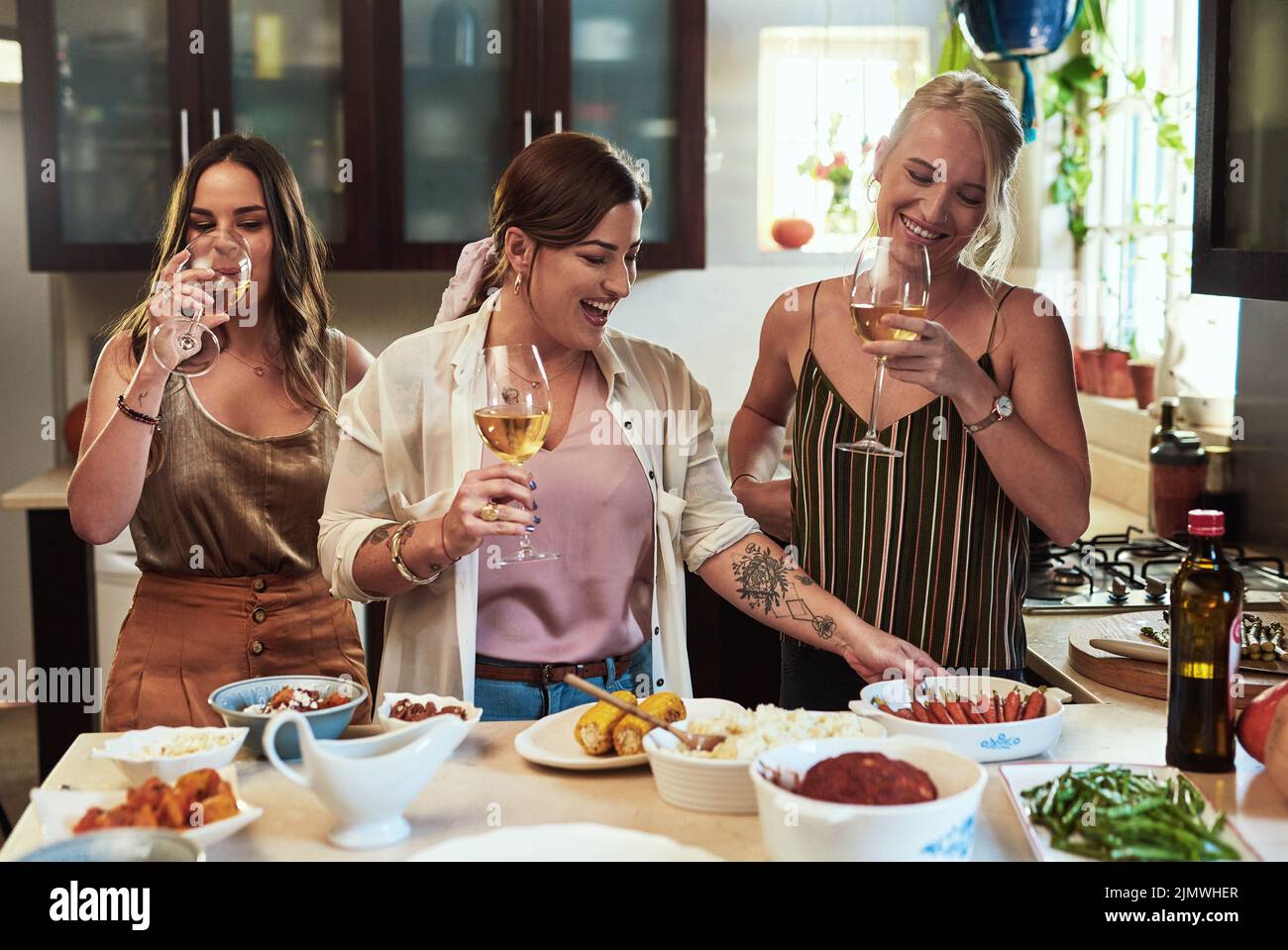Preparazione di una festa. Un gruppo di giovani amici allegri che preparano il cibo in cucina insieme mentre bevono vino a casa durante il giorno. Foto Stock