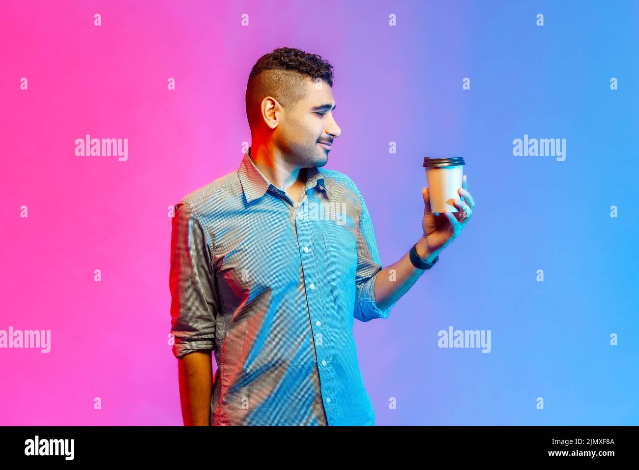 Ritratto di bel giovane uomo adulto in camicia che tiene una tazza di carta con caffè del mattino, guardando una bevanda calda, godendo. Studio interno girato isolato su colorato sfondo di luce al neon. Foto Stock