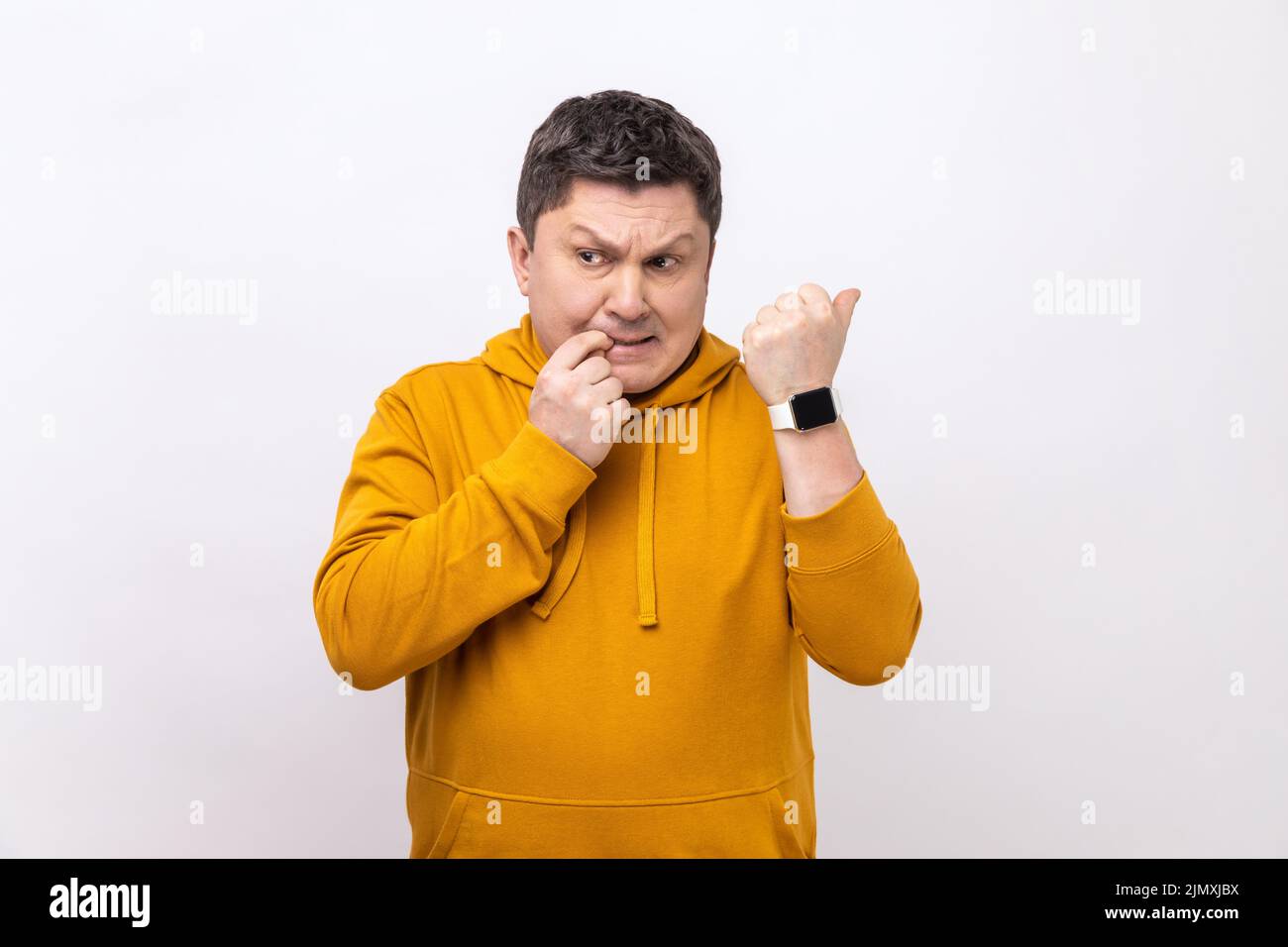 Uomo nervoso di mezza età mordendo le unghie delle dita e guardando il suo orologio da polso con ansioso, preoccupato per la scadenza, indossando la felpa con cappuccio urban style. Studio interno girato isolato su sfondo bianco. Foto Stock