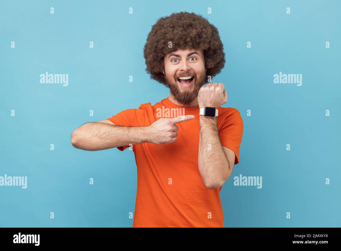 Ritratto di uomo sorridente positivo con acconciatura afroche indossa arancione T-shirt puntando orologio da polso ed esprimendo ottimismo sul tempo, non occupato. Studio interno girato isolato su sfondo blu. Foto Stock