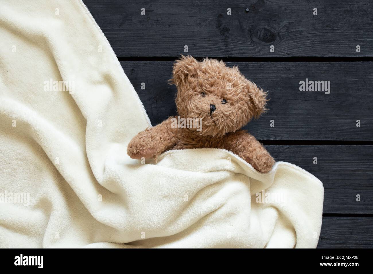 l'orso bruno di teddy si trova sotto una coperta bianca su uno sfondo di legno nero, bambino Foto Stock