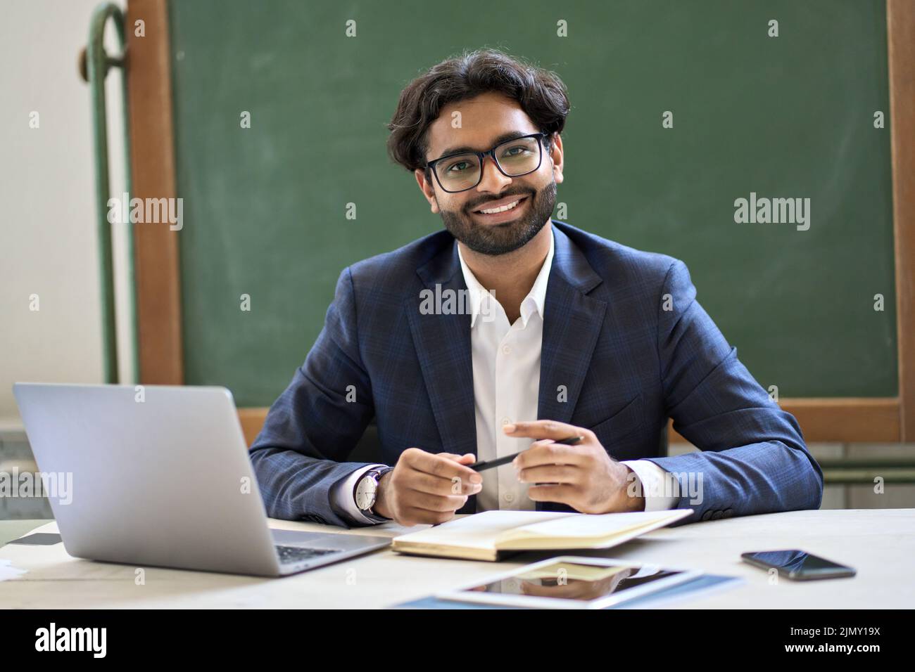 Buon giovane insegnante indiano di affari seduto al lavoro in classe. Verticale Foto Stock