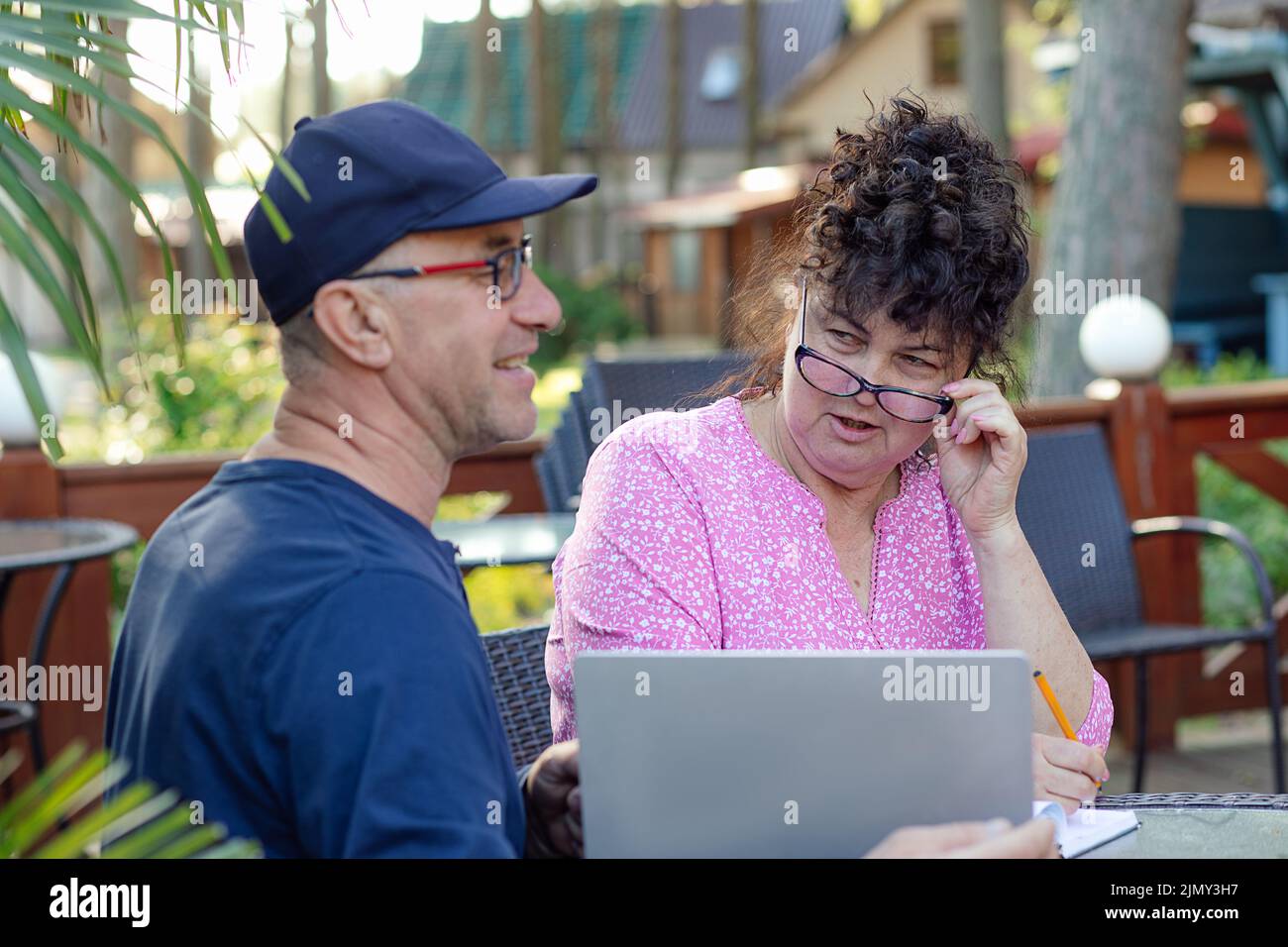Ritratto di coppia di mezza età. Donna curly che guarda sospetto a uomo sorridente seduto vicino a un computer portatile in un bar all'aperto. Foto Stock