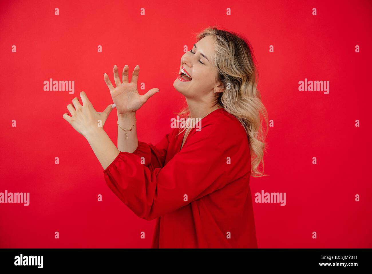 Ritratto vista laterale di donna sorridente in abito rosso che gioca su strumento invisibile con occhi chiusi isolato su sfondo rosso. Cantate e parlate Foto Stock