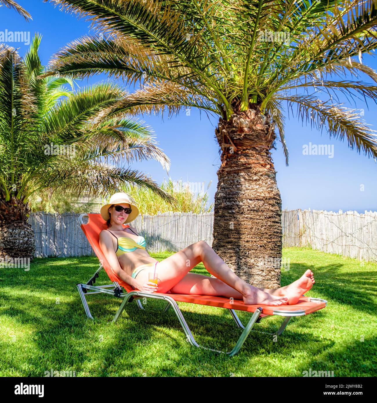 Donna con cappello sdraiato sul lettino in esotico resort tropicale sotto palme con bevanda in mano Foto Stock