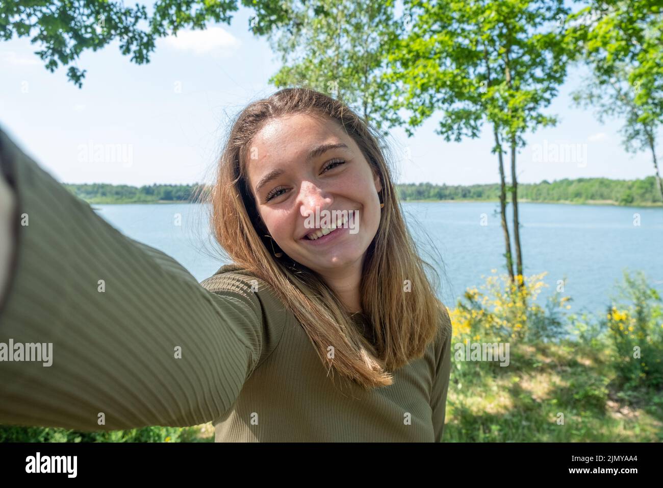 Bella giovane donna allegra che si divertiva al lago della foresta in una bella giornata, prendendo un selfie, sorridendo Foto Stock