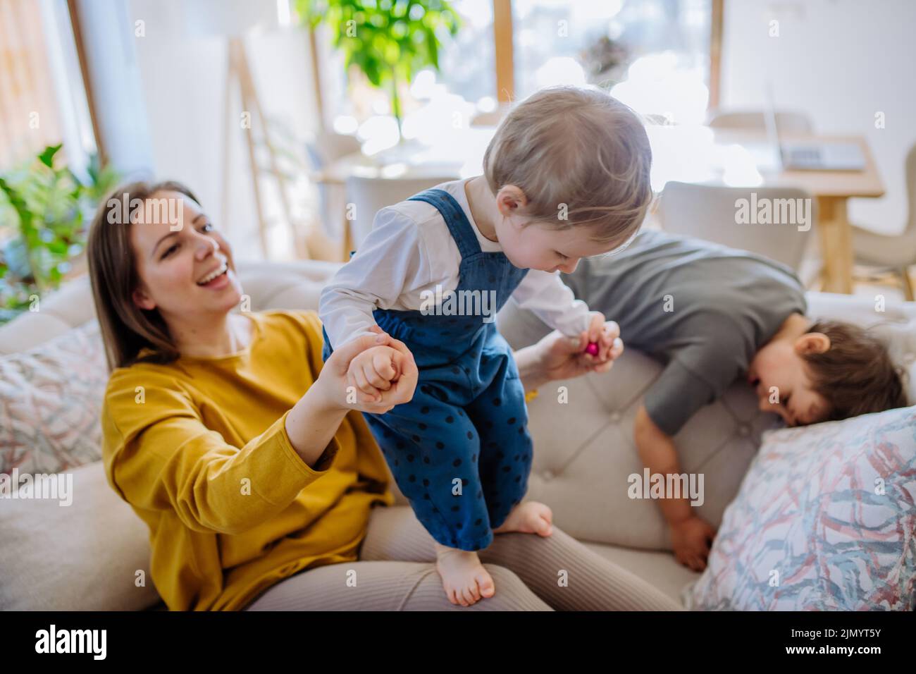 Giovane mamma allegra che gioca con i suoi bambini piccoli e si diverte quando li solleva sul divano. Foto Stock