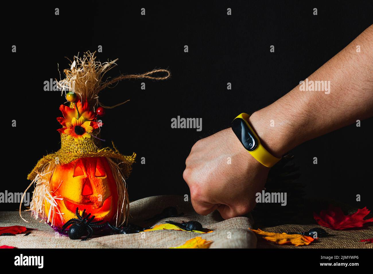 Braccio con un braccialetto per attività con fascia da polso gialla accanto a una zucca di Halloween adornata su sackcloth e circondata da ragni giocattolo e foglie cadute Foto Stock