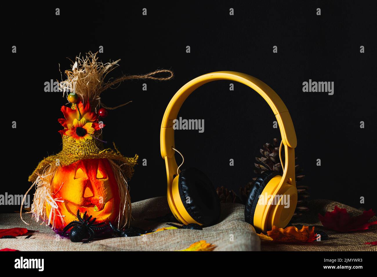 Cuffie wireless gialle in piedi accanto a una zucca ornata di Halloween su sackcloth e circondato da ragni giocattolo e foglie cadute accanto al cono di pino Foto Stock