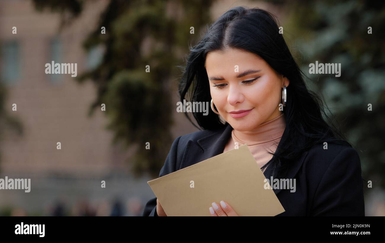 Ispanica giovane donna seduta su strada apre carta lettera leggere buone notizie bella ragazza felice sorridente femmina ha ottenuto sogno di carriera di crescita a Foto Stock