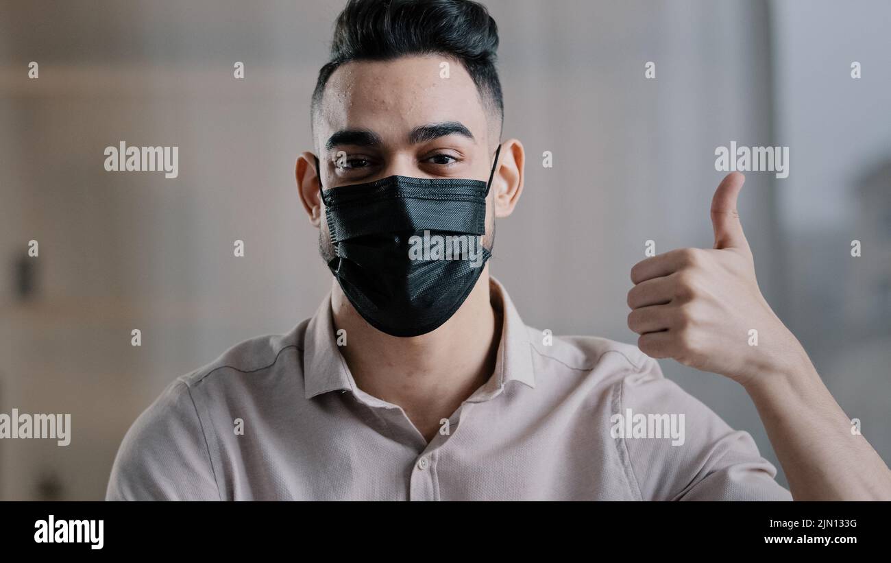 Etnia maschio studente uomo d'affari ispanico ragazzo in maschera medica proteggere da coronavirus focolaio in posa in casa mostrando pollici come gesto Foto Stock