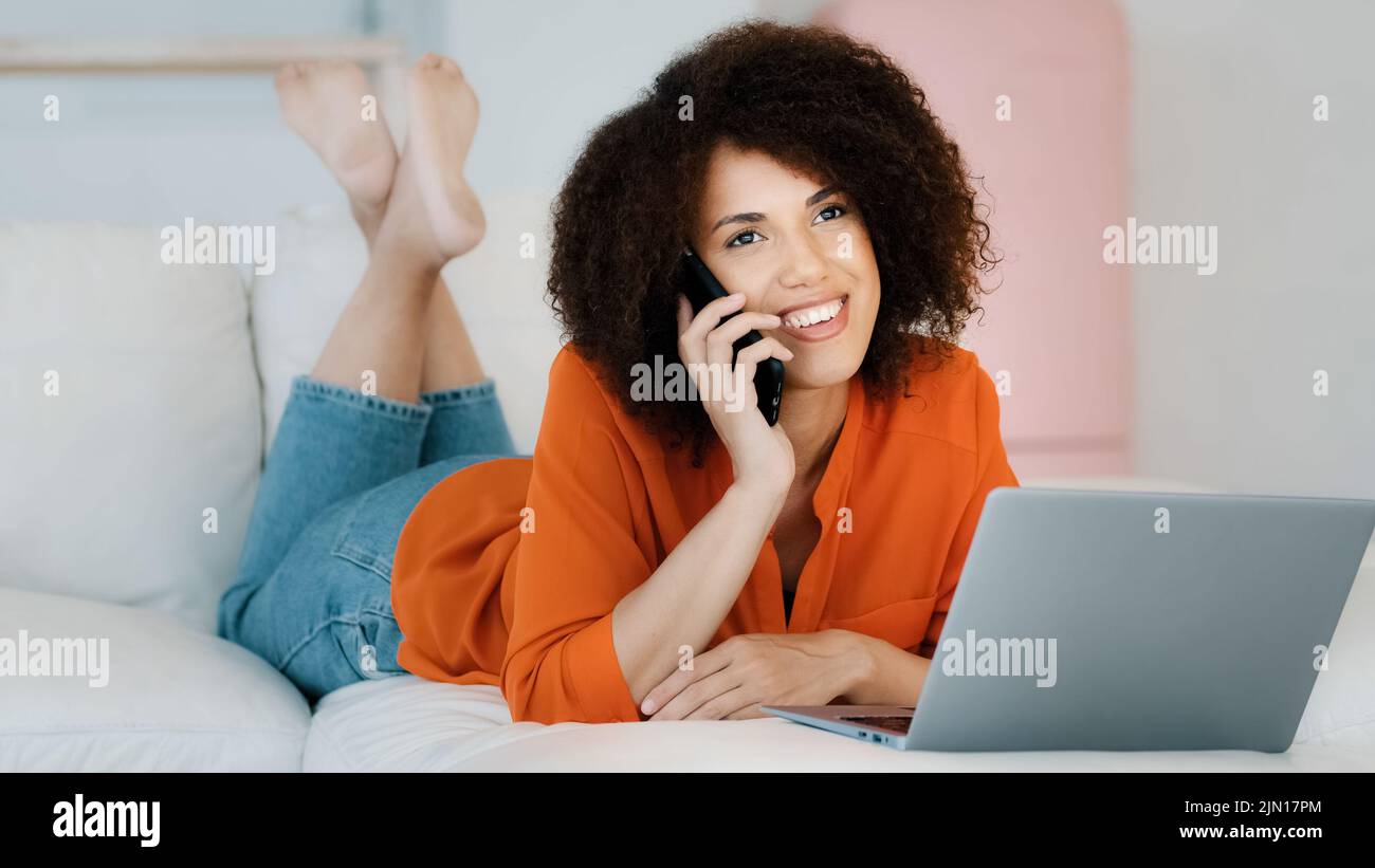 Sorridente ragazza afroamericana con capelli ricci sdraiati a casa a riposo parlando al telefono guardando lo schermo del computer portatile rilassante freelance donna fare Foto Stock