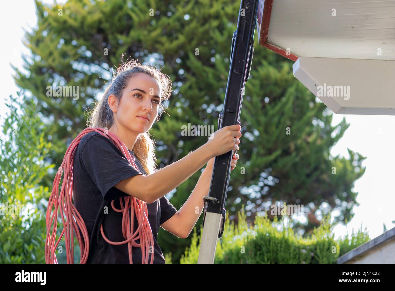 Ritratto di giovane bella donna che lavora in costruzione come elettricista con filo che sale la scala con retroilluminazione in giardino Foto Stock