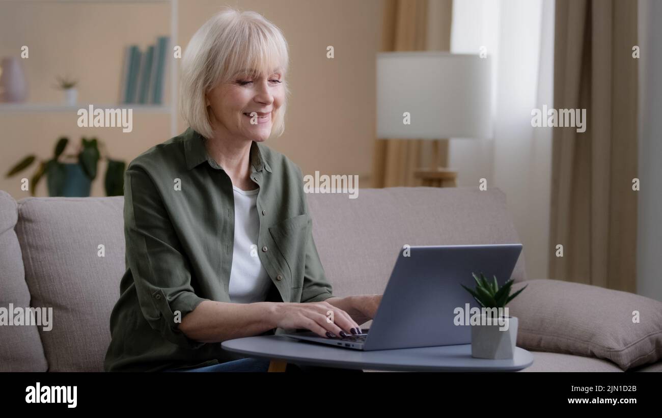 Vecchia femmina seduta sul divano a casa ufficio occupato a lavoro freelance con laptop anziano donna anziana digitando e-mail scrivere testo in computer web applicazione Foto Stock