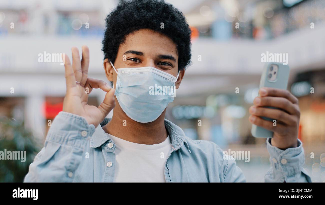 Ritratto africano dominicano ragazzo malato uomo in maschera medica protettiva con telefono che mostra ok gesto alla fotocamera raccomanda buona app medica felice con Foto Stock