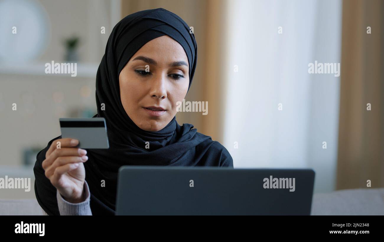 Sorridendo cliente della ragazza islamica in hijab seduta sul divano fare ordine in linea usare il laptop inserire il numero della carta di credito per la transazione felice donna musulmana pagare Foto Stock