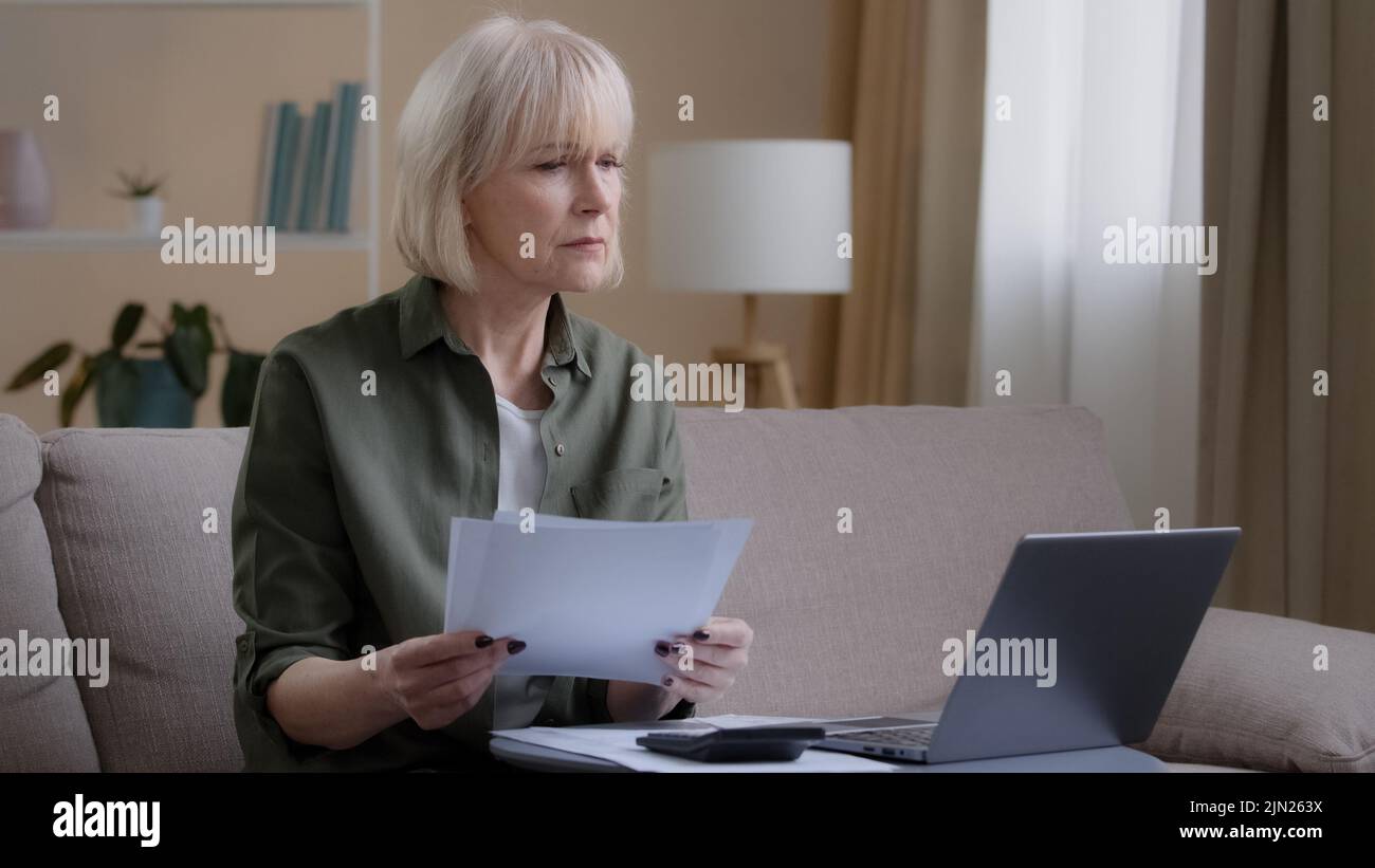 Occupato 50s casalinga padrone di casa fa la donna bilancio controlla il reddito mensile controllando le note della carta usando il laptop dell'app. Donna d'affari di mezza età seduta sopra Foto Stock
