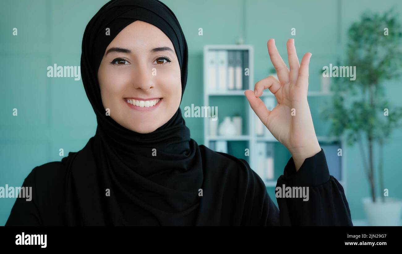 Ritratto al coperto musulmana ragazza islamica sorride toothy largo alla macchina fotografica mostrando mano ok segno. Studentessa araba mostra tutto il bel gesto di successo Foto Stock