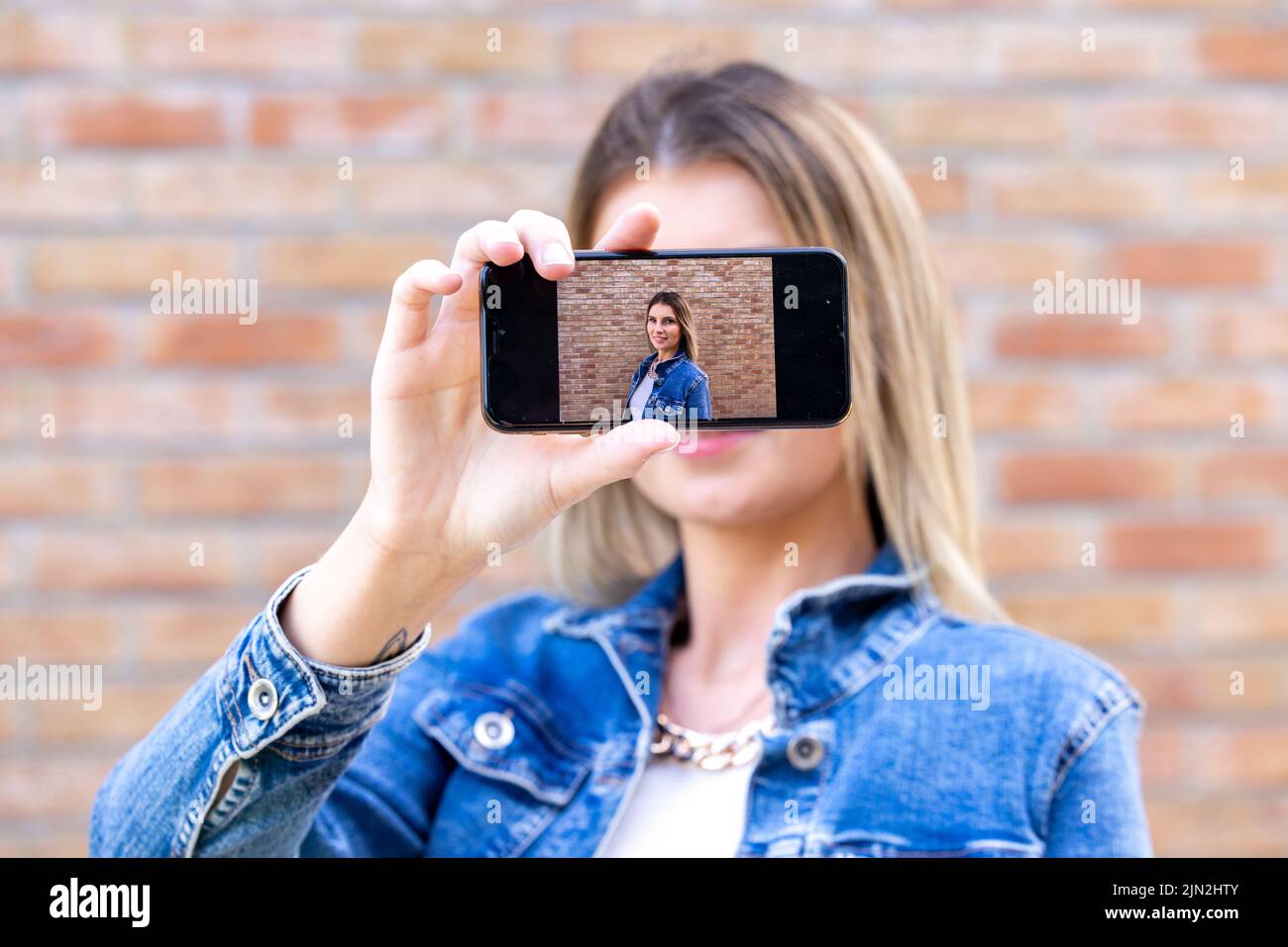 Foto di allegro carino affascinante bella bella ragazza bionda attraente, con pelle pallida mostrando selfie con il suo telefono tenuto nelle sue mani indossare giacca jeans isolato su sfondo rosso mattone colore. Foto di alta qualità Foto Stock