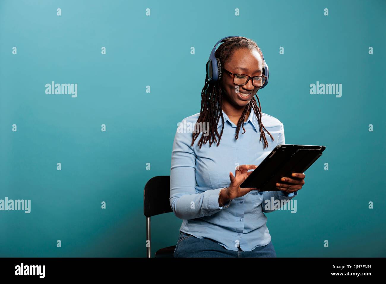 Bella giovane donna felice con un moderno dispositivo tablet touchscreen sorridente mentre navighi su Internet. Persona gioiosa con dispositivo elettronico wireless che naviga le pagine web mentre si siede su sfondo blu. Foto Stock