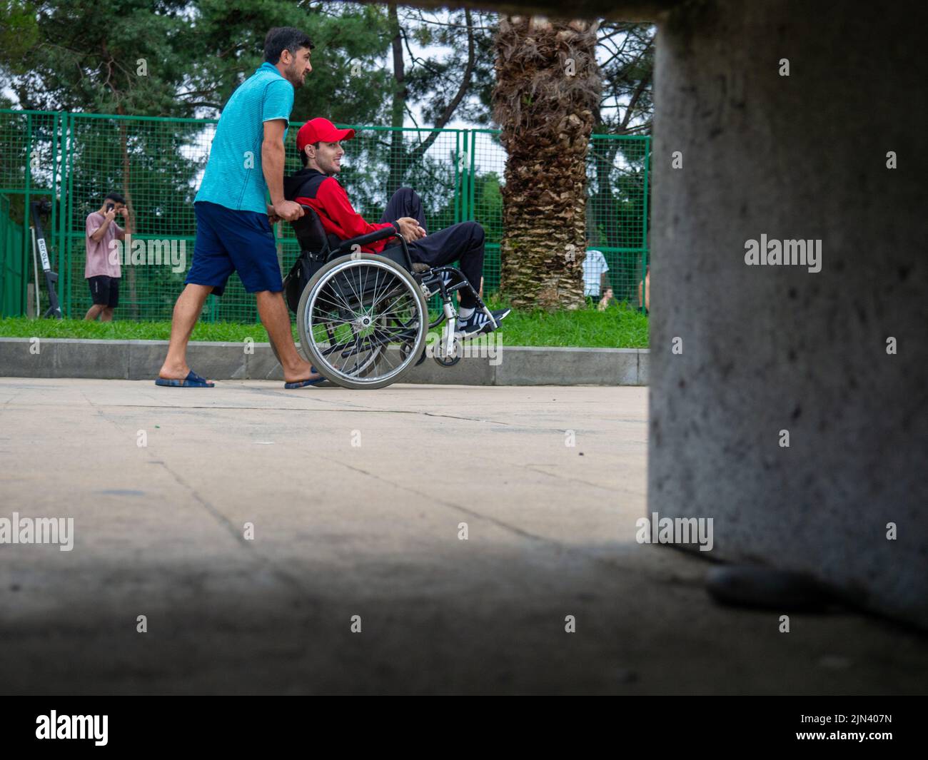Batumi, Georgia. 07.19.2022 Una persona in sedia a rotelle viene fatta rotolare nel parco da un'altra persona. Passeggiata nel parco. La vita delle persone. Foto Stock
