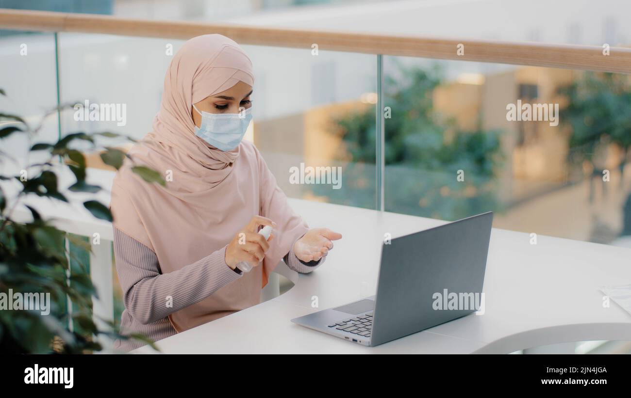 Divertente donna araba seduta in luogo pubblico usa antisettico spruzzi intorno in aria su laptop copre faccia con maschera medica ragazza musulmana paura di germi Foto Stock