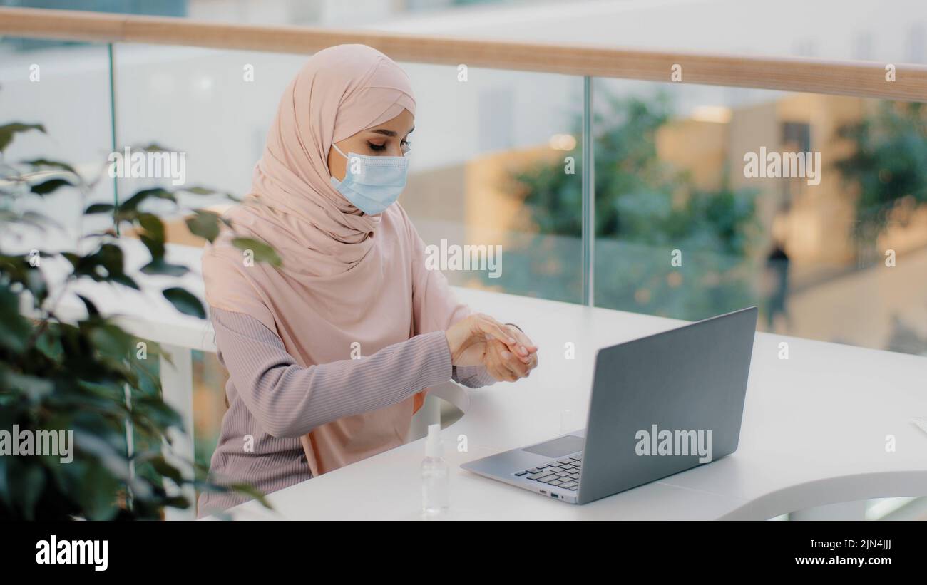 Divertente donna araba seduta in luogo pubblico usa antisettico spruzzi intorno in aria su laptop copre faccia con maschera medica ragazza musulmana paura di germi Foto Stock
