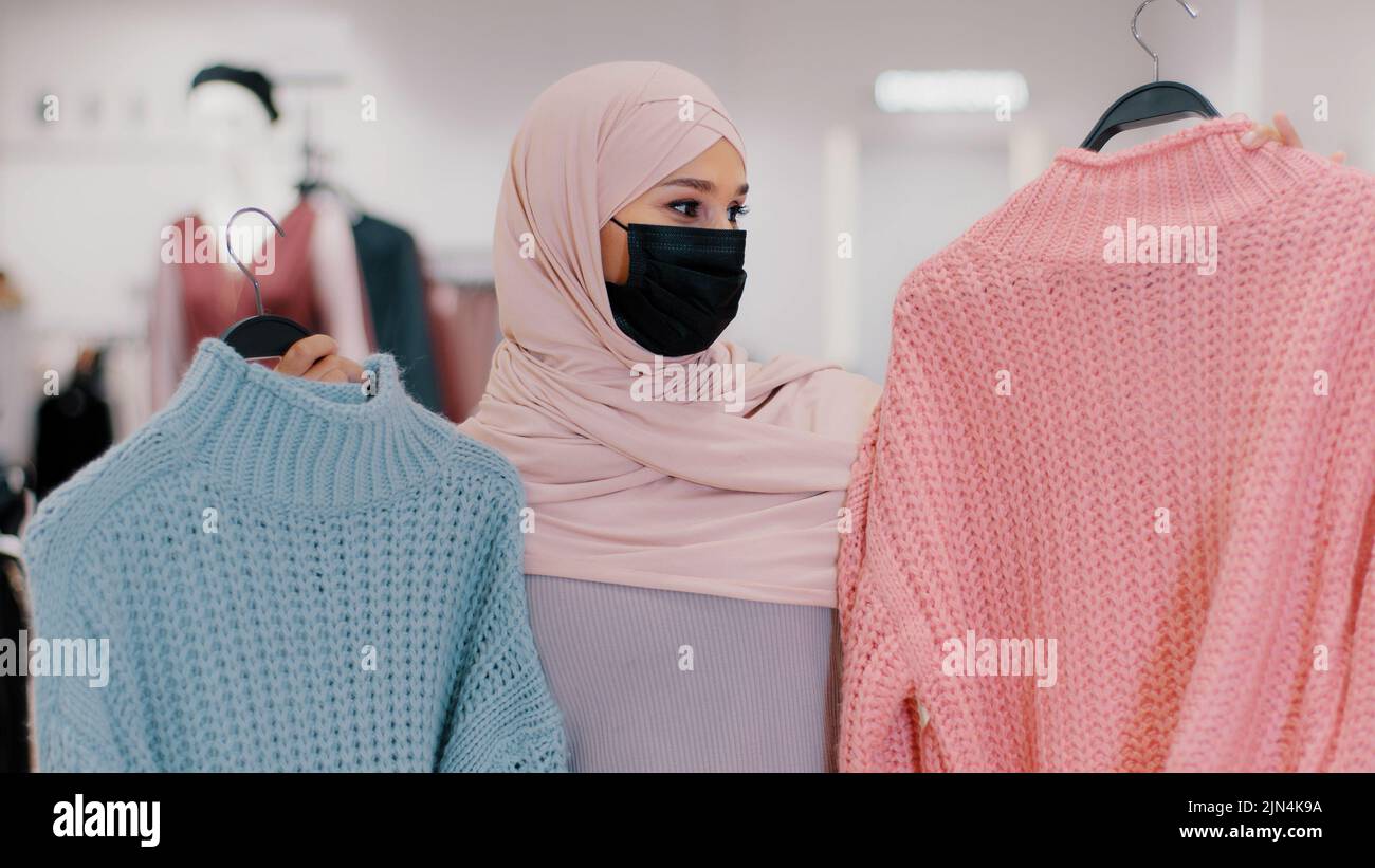 Donna musulmana giovane in maschera medica protettiva durante la quarantena pandemica ragazza islamica in hijab venditore in negozio di abbigliamento offre maglioni in vendita Foto Stock