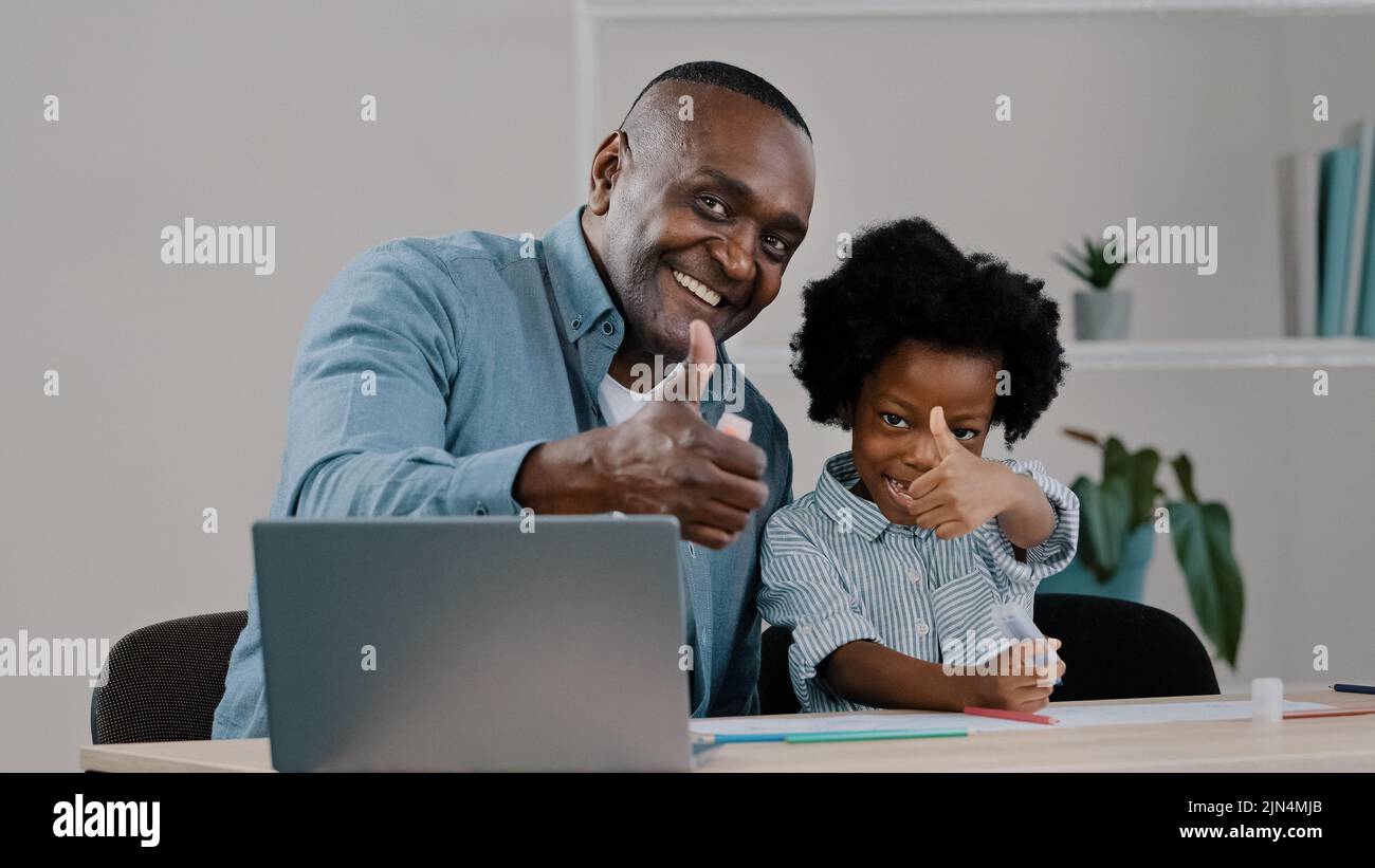 Felice afroamericano maturo padre prendersi cura sorride aiutando bambina scolaretta con studio lavoro mostrando pollici su gesto di approvazione utilizzando Foto Stock